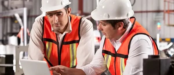 Two production planners review a laptop on the factory floor to build a stronger manufacturing production schedule with APS software.